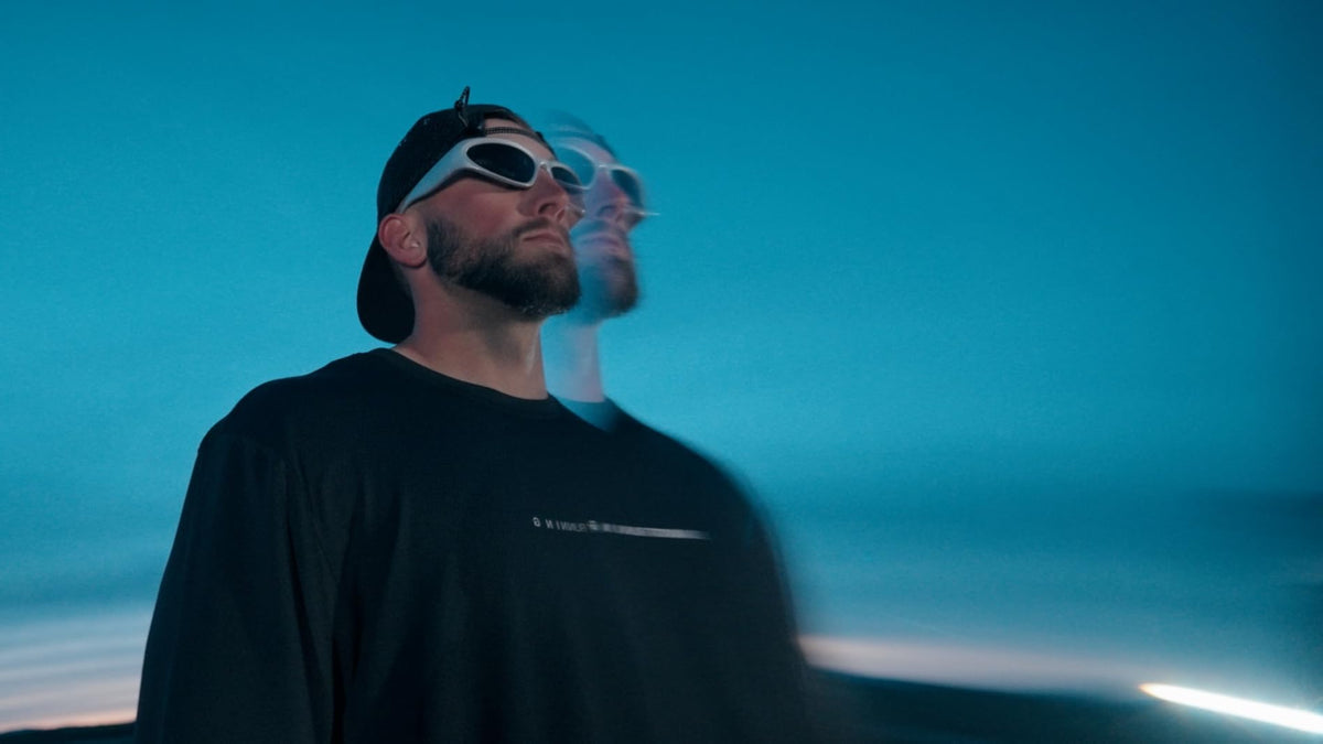 A man wearing a black long-sleeve t-shirt, a black baseball cap, and white-framed sunglasses, captured in a motion-blurry style against a blue twilight sky. A faded duplicate of his face and upper body appears behind him.