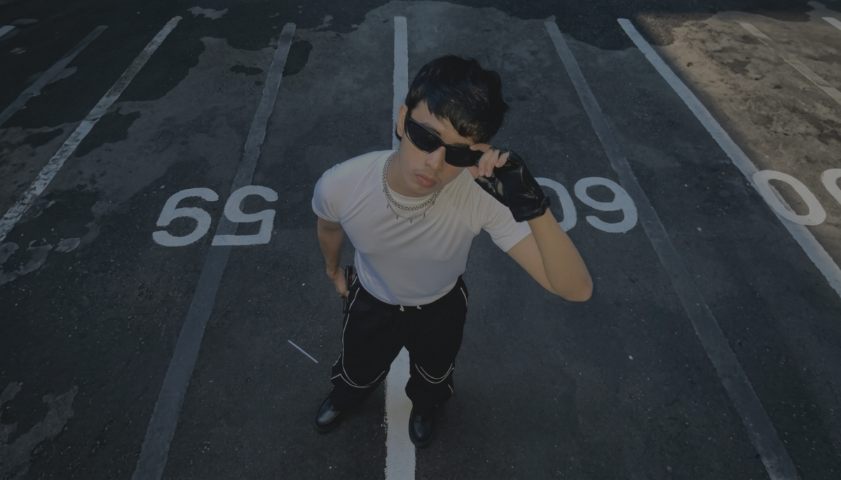 A man wearing a white t-shirt, black pants, black sunglasses, and black gloves stands in a parking lot, looking up towards the camera.