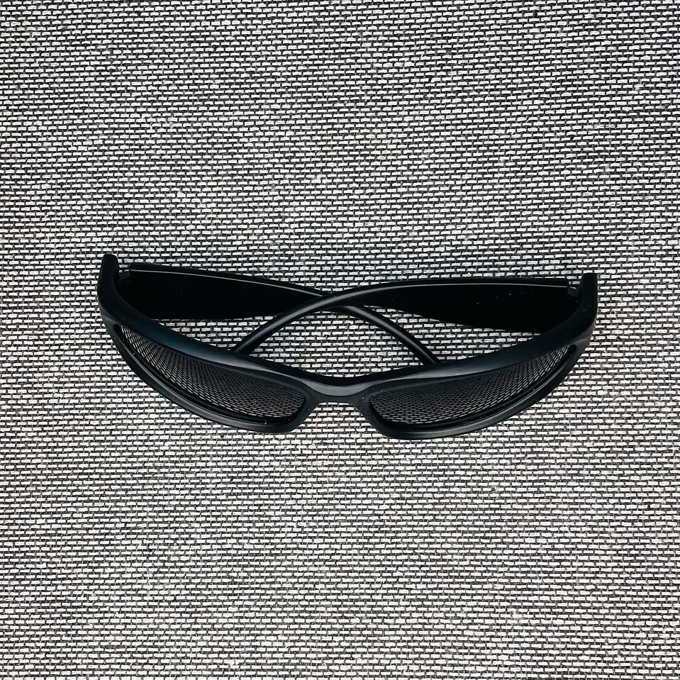 Black safety glasses with a curved design and mesh lenses, resting on a textured gray woven fabric background.