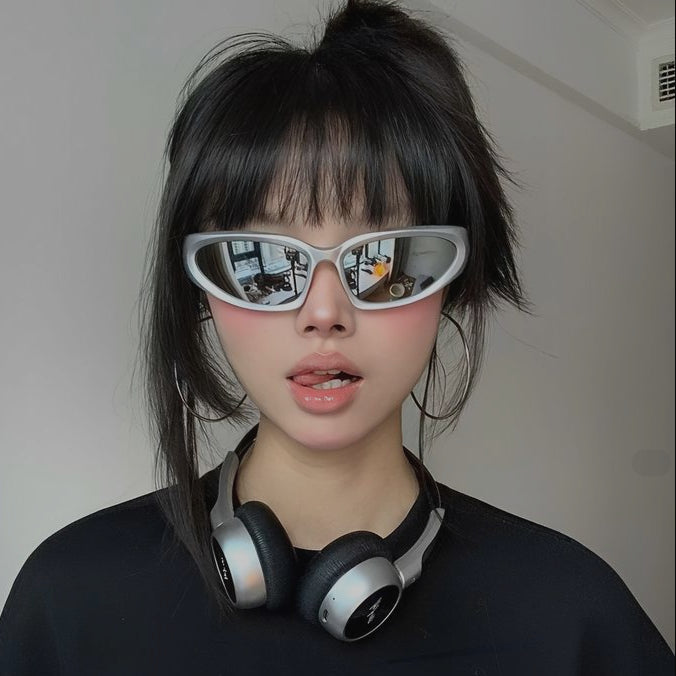 A person with dark hair wearing cat-eye sunglasses with silver frames and black-and-silver over-ear headphones around their neck, against a plain indoor wall.